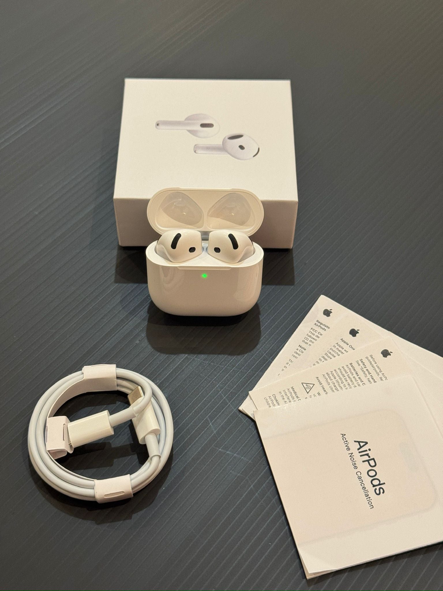 Beige wireless earbuds in charging case with box, cable, and manual on dark surface